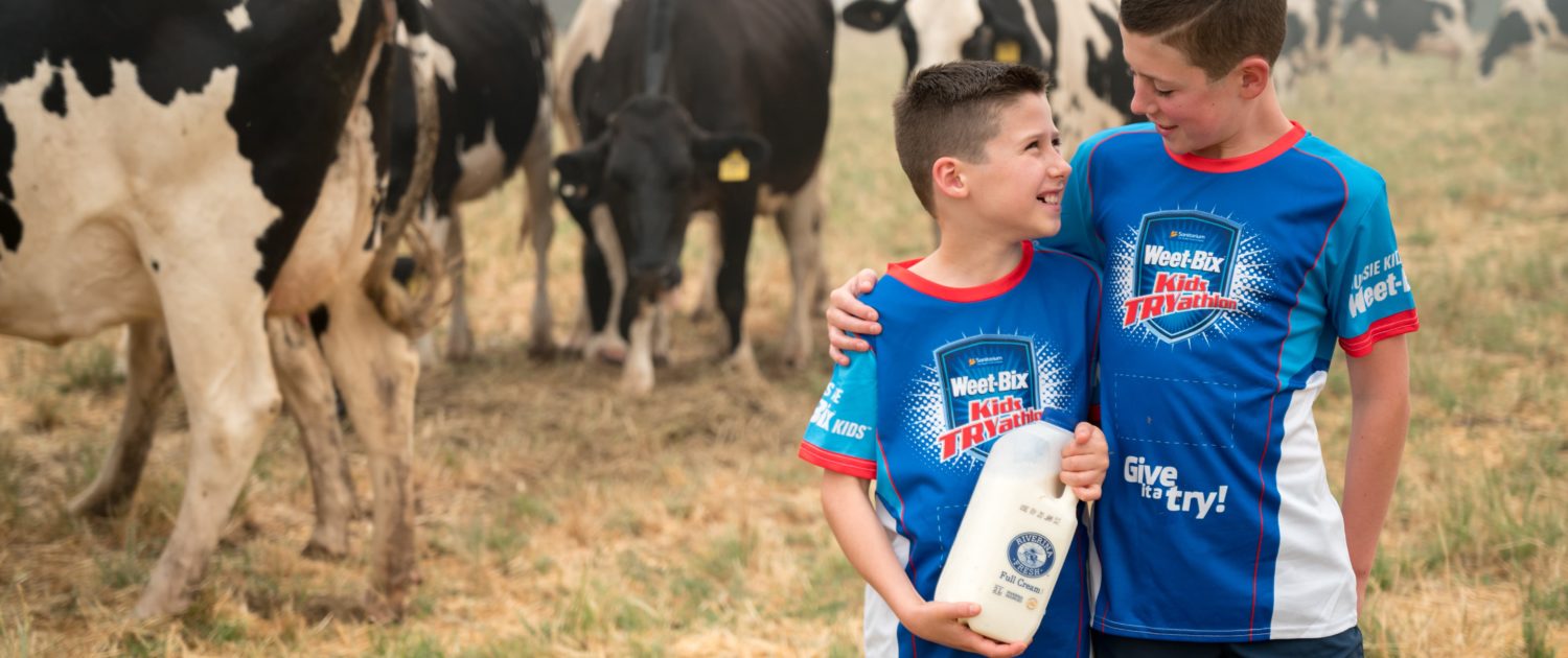 WeetBix TryAthalon-min Two children standing in front of cows holding Riverina Fresh milk wearing Weet-Bix TRYathalon shirts